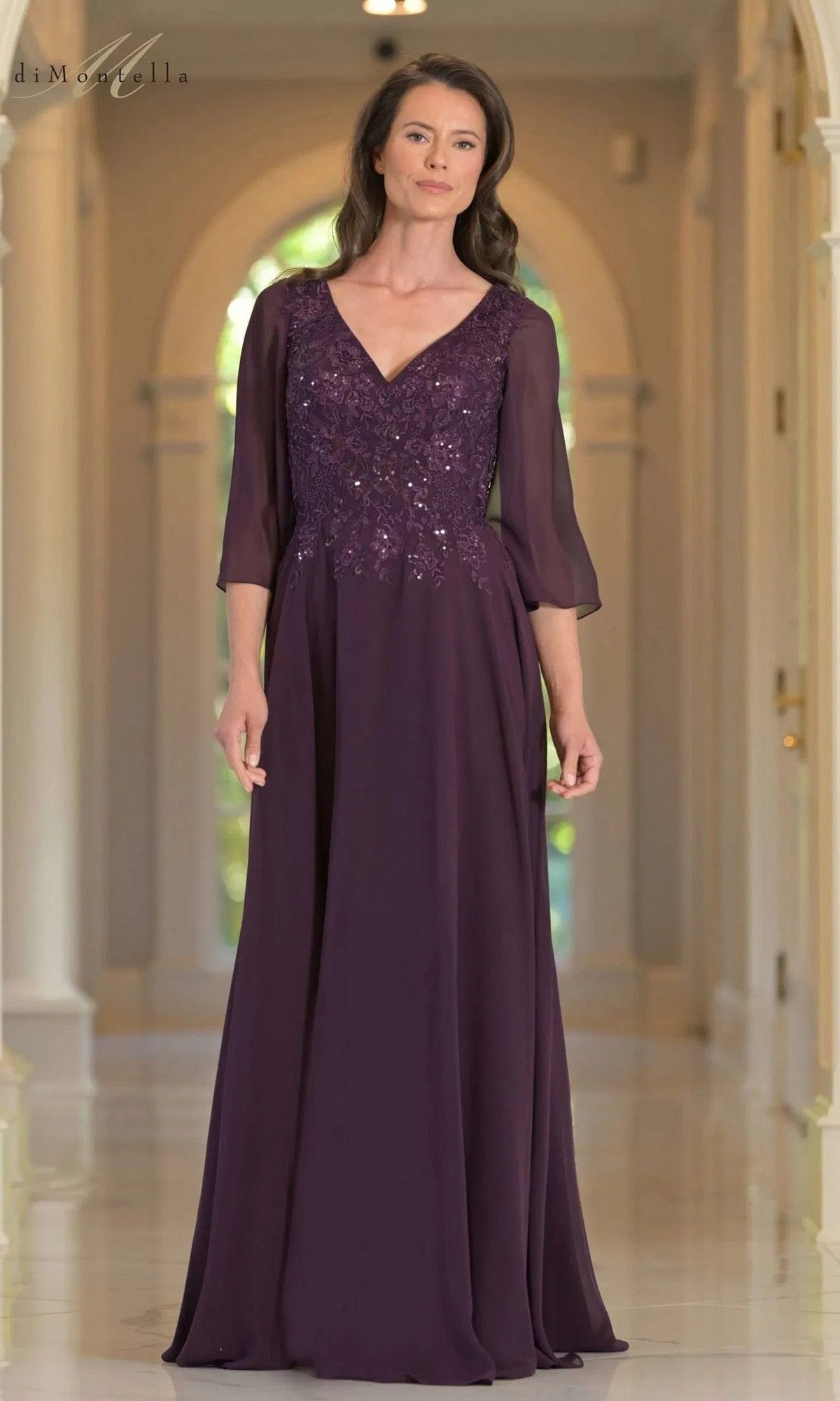 A woman stands in a hallway wearing the Rina Di Montella RD3241 long formal dress in eggplant, featuring sheer sleeves and an elegantly sequin-embellished, floral-embroidered bodice.